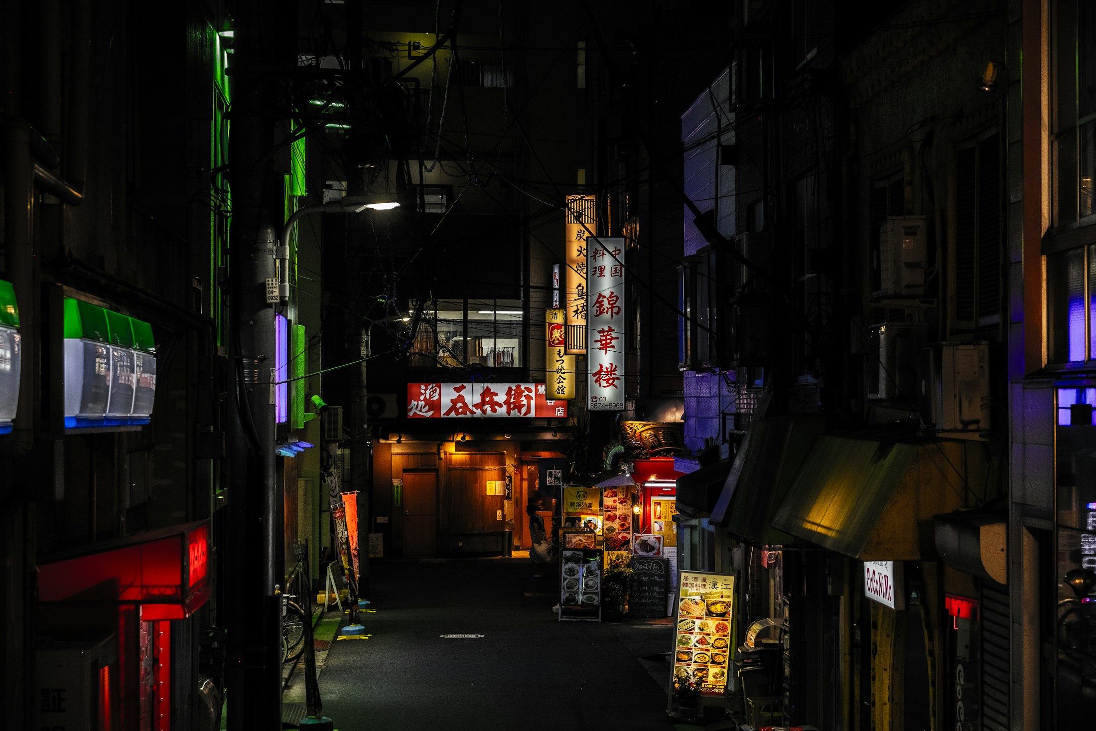 A narrow city alley at night, crossed by wires and lit with green and red signs.